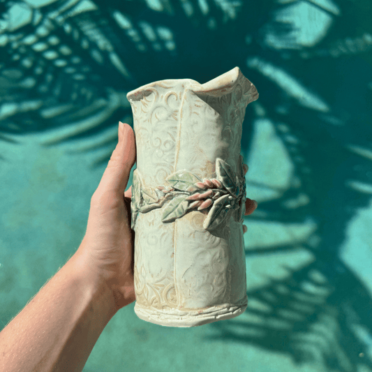 Hand holding a decorative ceramic vase with leaf designs against a green leafy background. Hand Made Sunshine Coast.