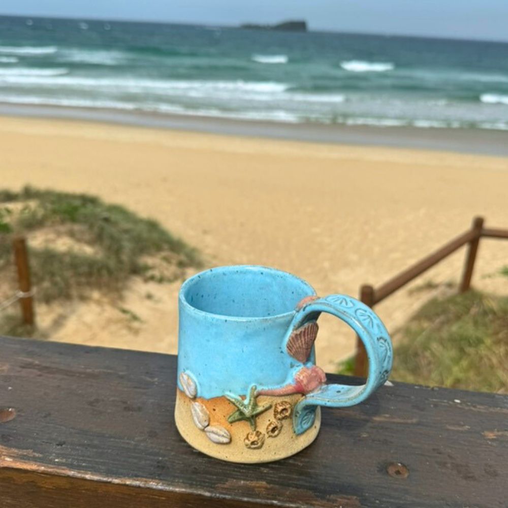 Blue ceramic mug with beach-themed design on a wooden surface by the ocean.