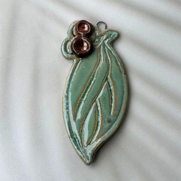 Ceramic leaf-shaped ornament with two small copper-colored beads on a light background