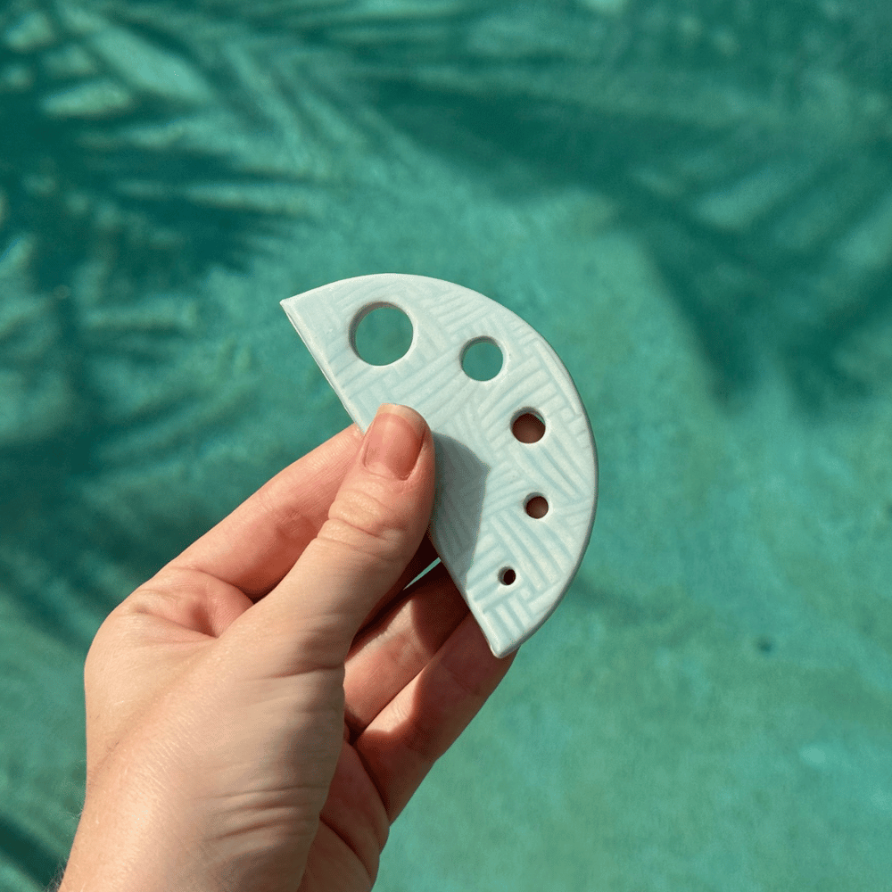 Hand holding a light blue ceramic herb shredder with holes against a teal background