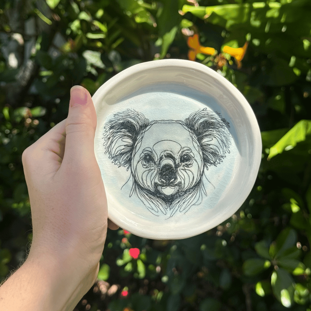 Hand holding a white dish with a black and white illustration of a koala on a green leafy background