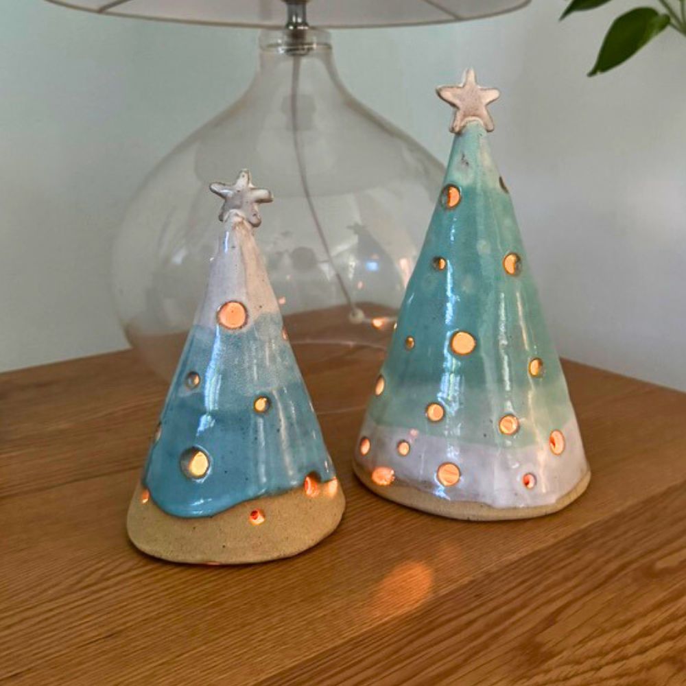 Two ceramic Christmas tree luminary cones on a wooden surface with a lamp in the background.
