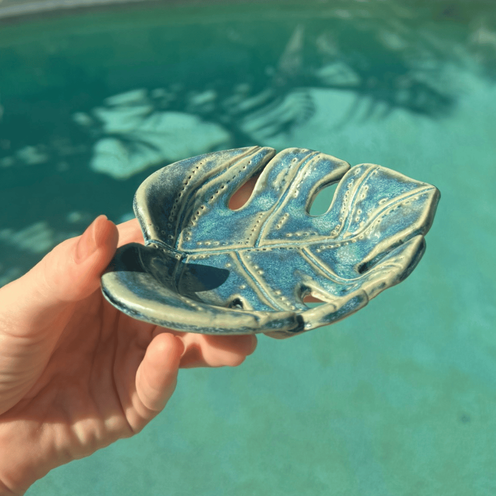 Hand holding a blue monstera ceramic leaf-shaped dish against a pool background