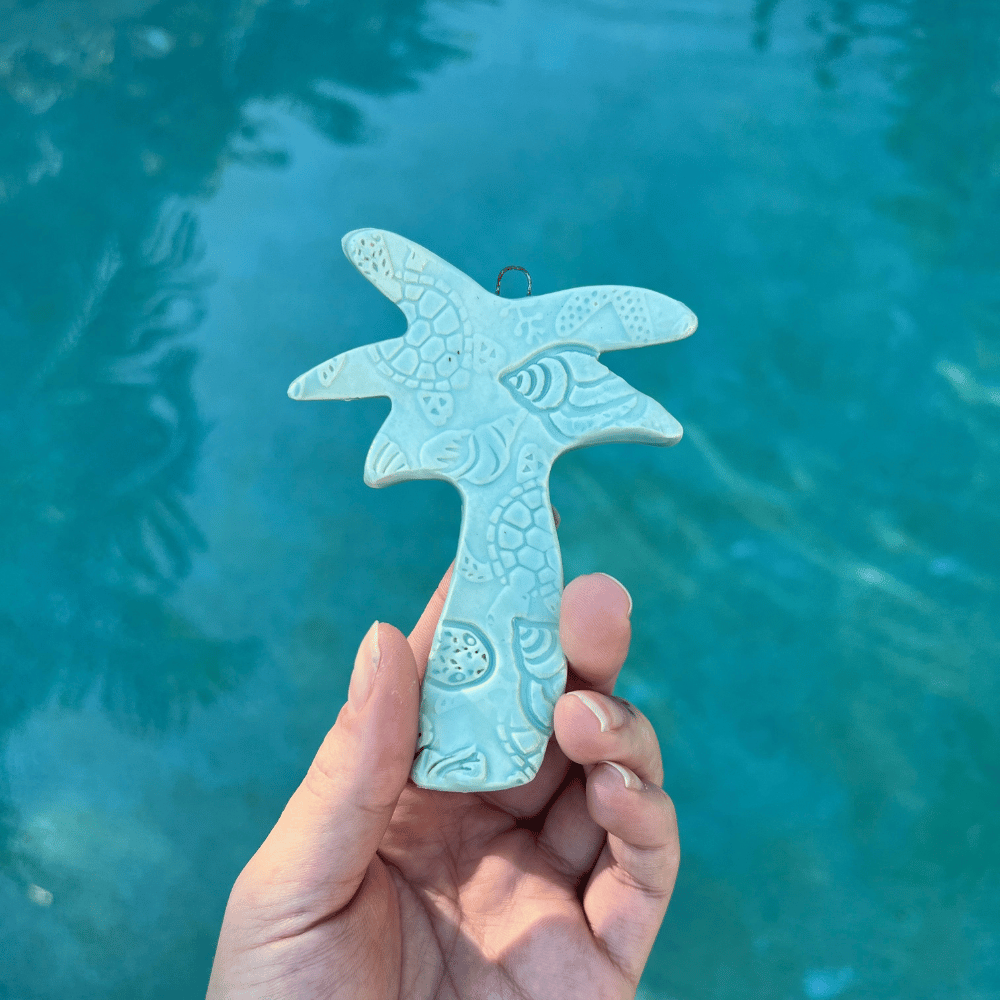Hand holding a light blue palm tree-shaped ornament against a teal water background. Sunshine Pot-Tree, Sunshine Coast, Queensland, Australia