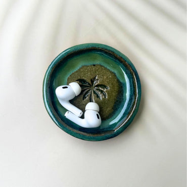 White wireless earbuds on a green ceramic dish with a palm tree design on a white background