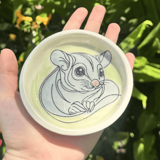 Hand holding a trinket dish with a sugar glider illustration against a green leafy background
