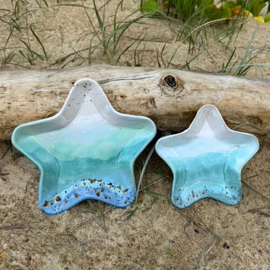 Two star-shaped ceramic bowls with a gradient blue color on a sandy surface with driftwood and grass.