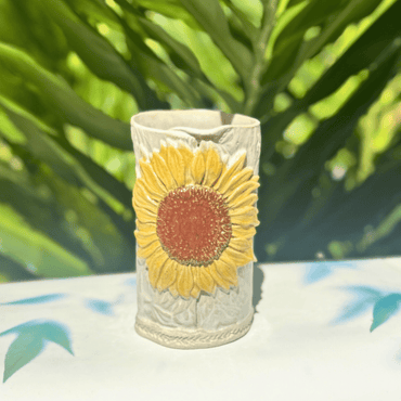 Ceramic vase with sunflower design on a white surface with green foliage in the background