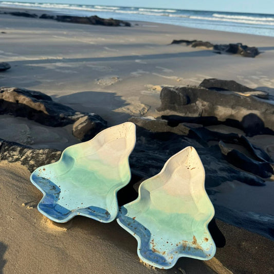 Two ceramic xmas tree plates on a sandy beach with rocks and ocean in the background.