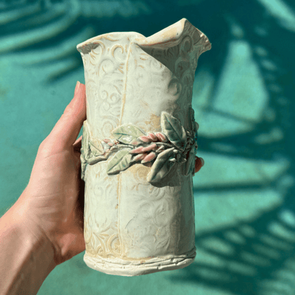 Hand holding a decorative ceramic vase with leaf designs against a teal background. Hand Made Sunshine Coast.