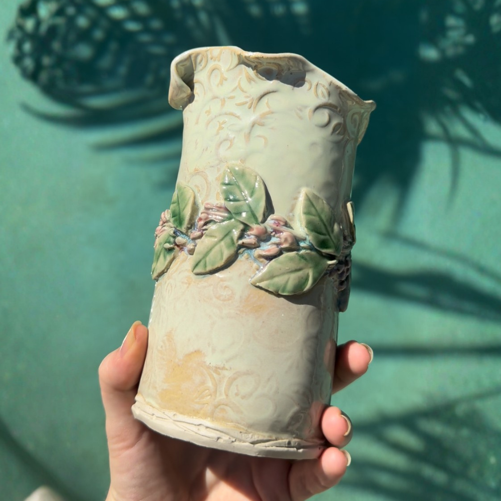 Hand holding a ceramic vase with leaf designs against a green background