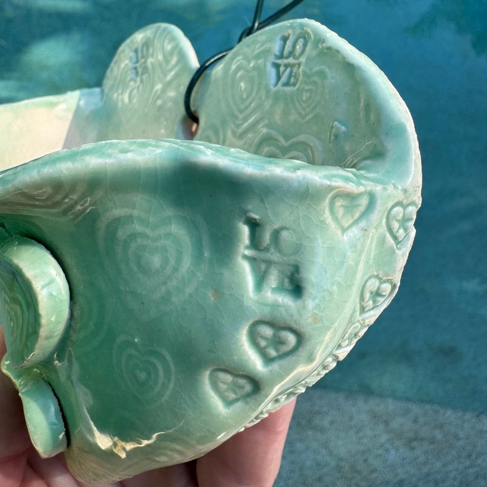 Green ceramic heart-shaped wall planter with 'LOVE' and heart patterns, held against a blue background.