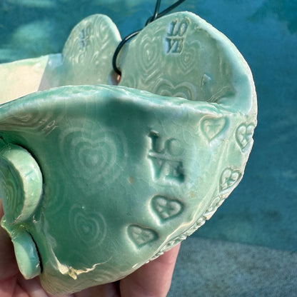 Green ceramic heart-shaped wall planter with 'LOVE' and heart patterns, held against a blue background.