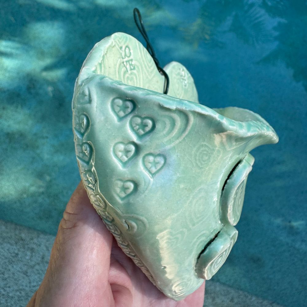 Hand holding a green ceramic heart-shaped wall planter with heart patterns against a blue water background