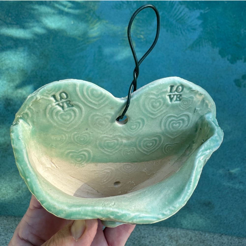 Hand holding a green ceramic heart-shaped wall planter with heart patterns against a blue water background