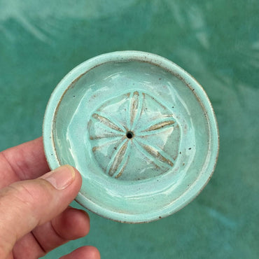 Hand holding a small turquoise ceramic incense holder with sand dollar center against a teal background
