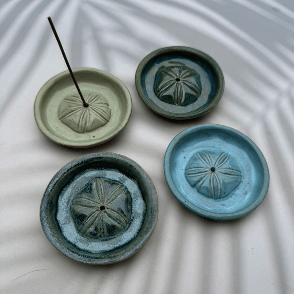 Four ceramic incense holders in different shades of green with sand dollar design on a textured white surface.