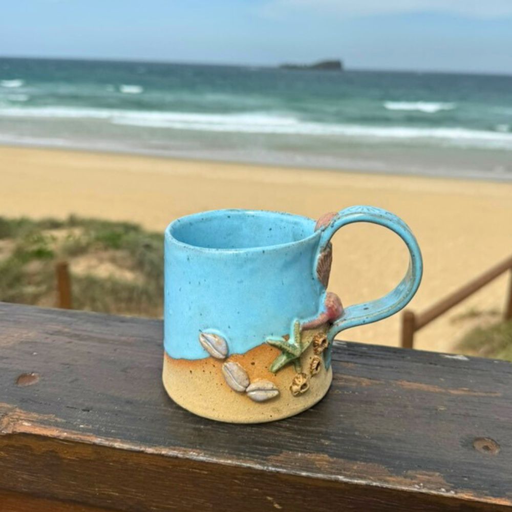 Blue ceramic mug with beach-themed decorations on a wooden surface with a beach in the background