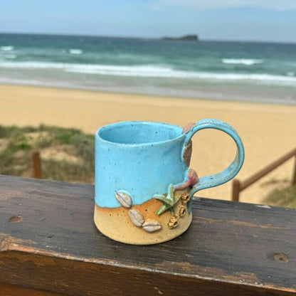 Blue ceramic mug with beach-themed decorations on a wooden surface with a beach in the background
