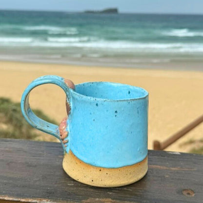 Blue ceramic mug on a wooden surface with a beach and ocean in the background