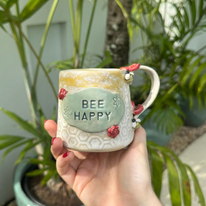 Hand holding a mug with 'BEE HAPPY' text against a plant background