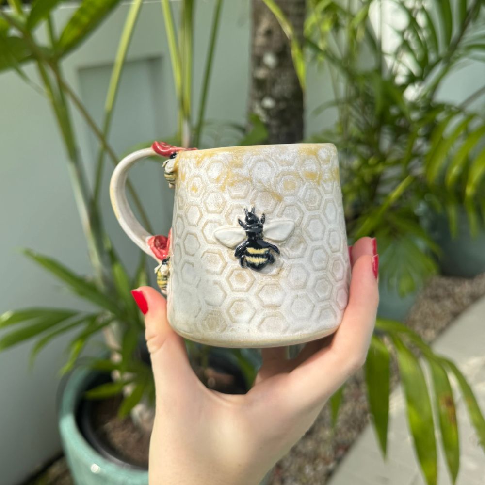 Hand holding a decorative mug with a bee and honeycomb design, surrounded by plants.