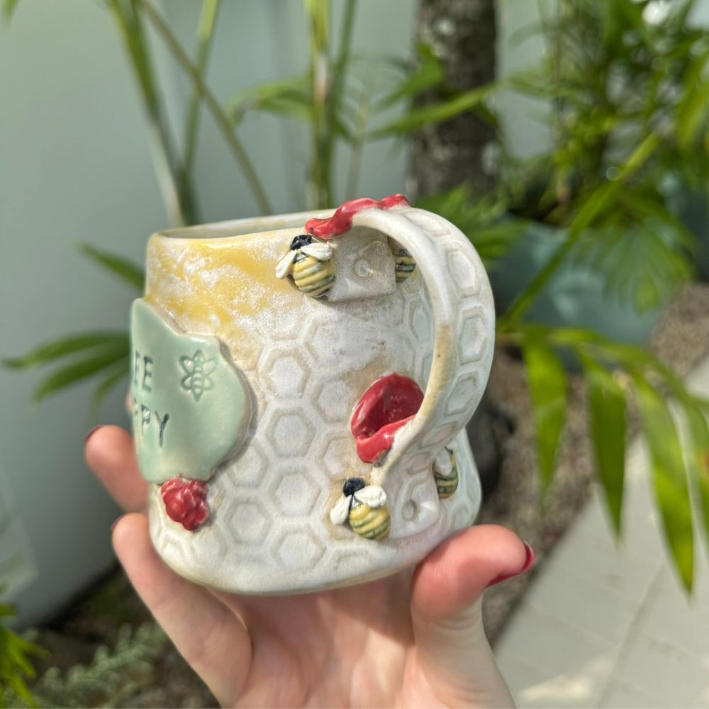 Ceramic mug with bee and honeycomb design held in a hand, outdoor background