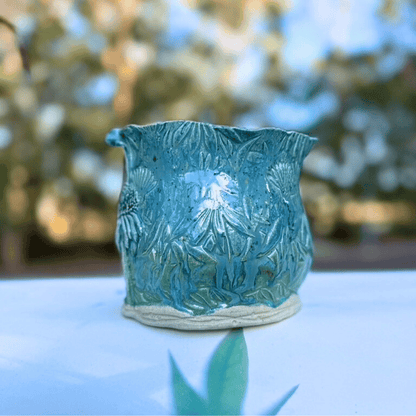 Teal ceramic planter with floral design on a white surface with blurred greenery in the background