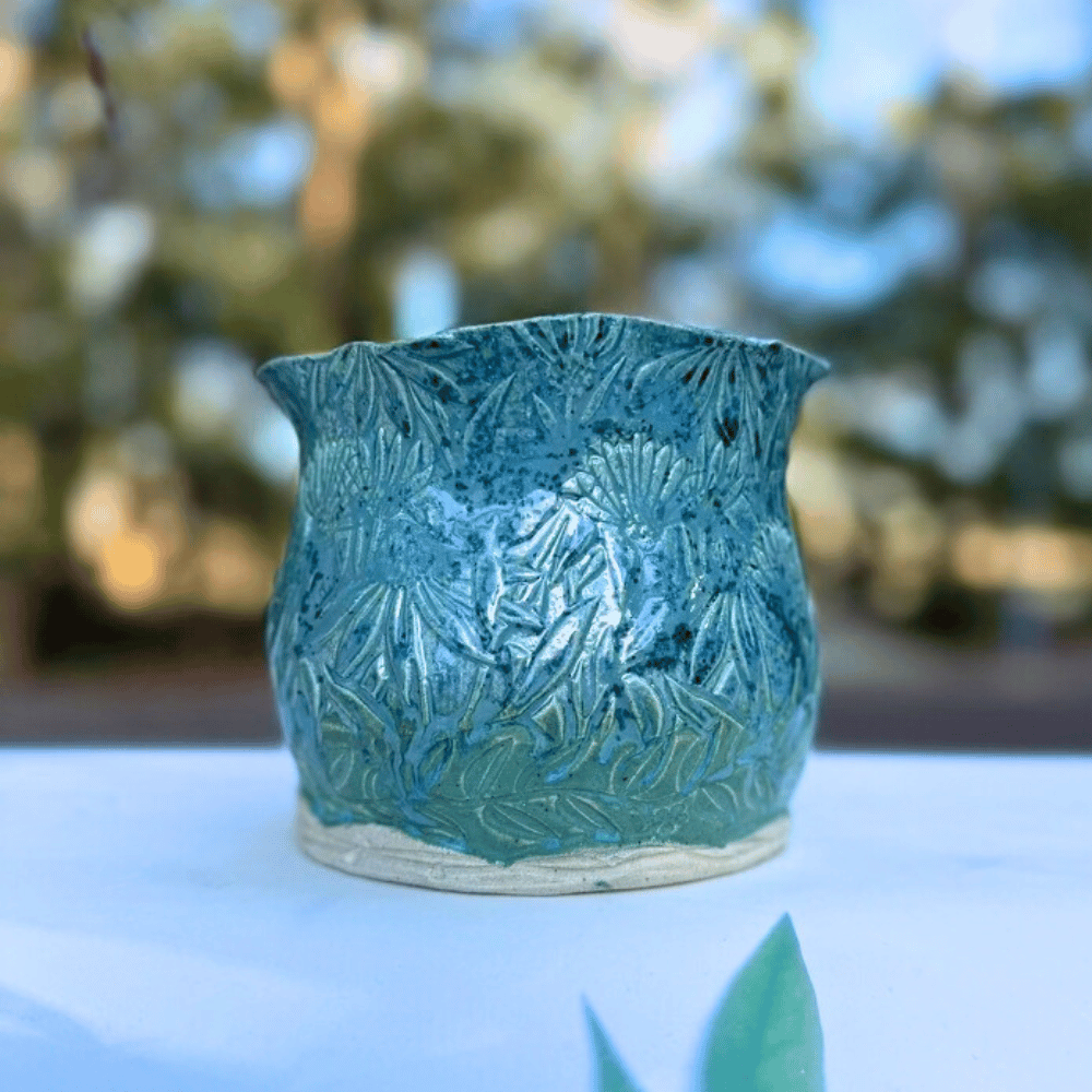 Teal ceramic planter with floral design on a white surface with blurred greenery in the background