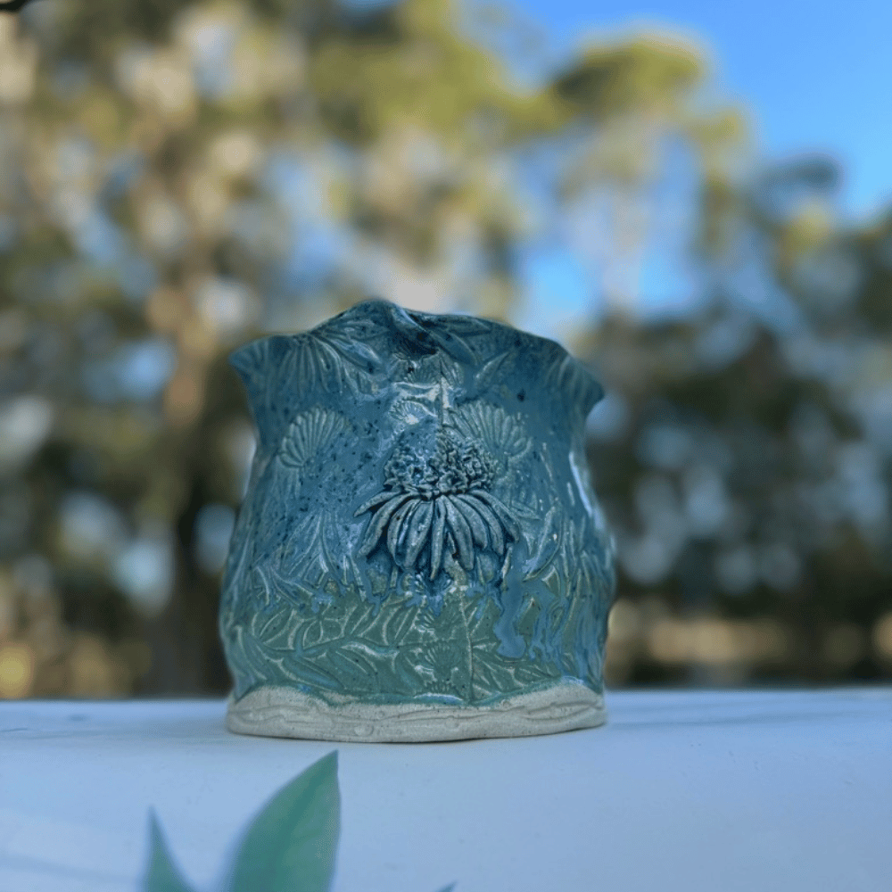 Teal ceramic planter with floral design on a white surface with blurred greenery in the background