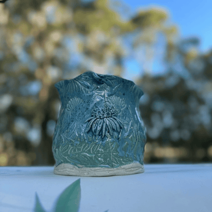 Teal ceramic planter with floral design on a white surface with blurred greenery in the background