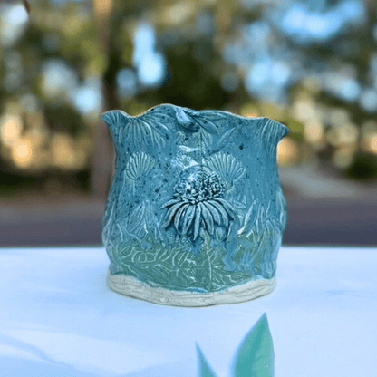 Teal ceramic planter with floral design on a white surface with blurred greenery in the background