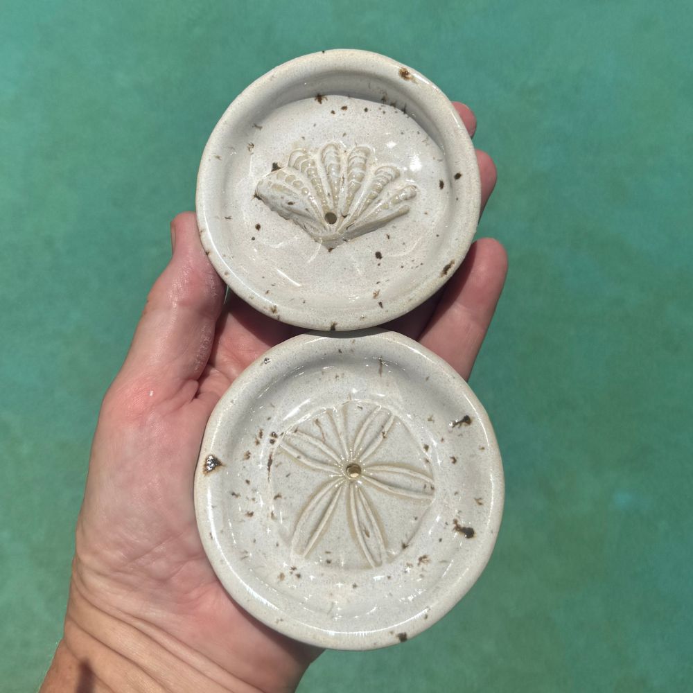 Two ceramic incense holders with shell designs held against a teal background