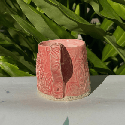 Pink ceramic vase with textured design on a white surface with green leaves in the background