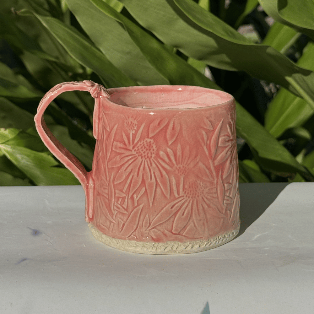 Pink ceramic handmade mug with floral patterns on a white surface with green plants in the background
