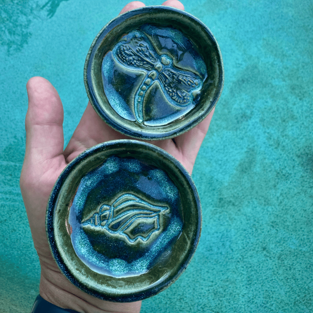 Two ceramic trinket with dragonfly designs held against a teal background