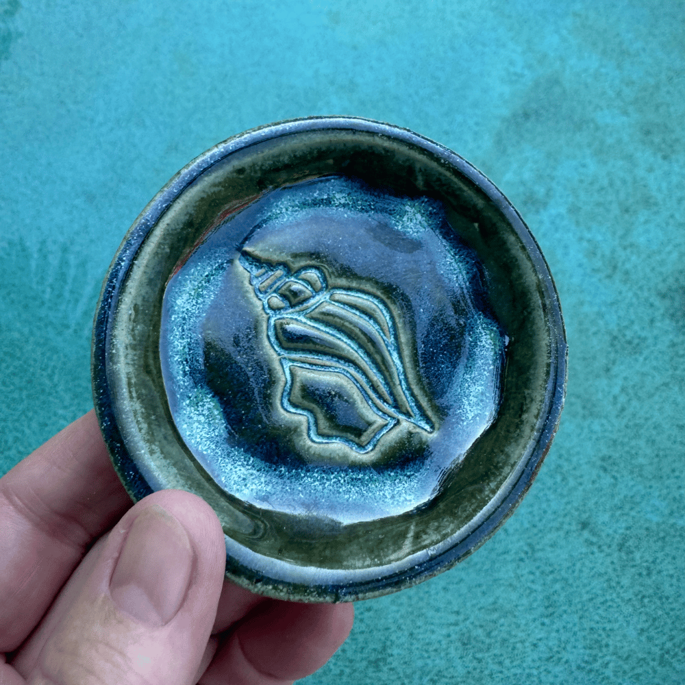 Hand holding a ceramic trinket dish with a shell design against a teal background
