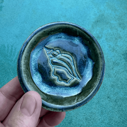 Hand holding a ceramic trinket dish with a shell design against a teal background