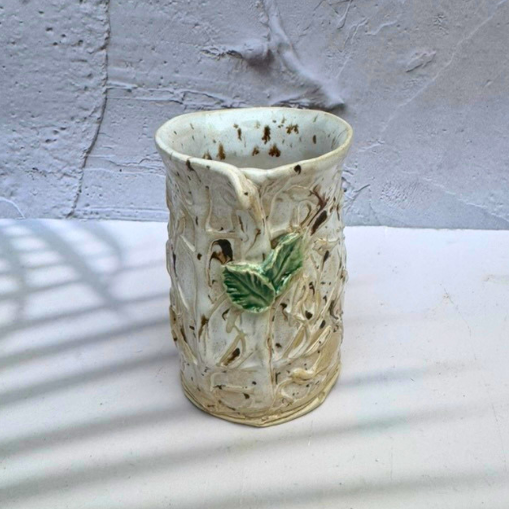 A handmade ceramic vase with a textured, organic leafy pattern in cream and brown glazes, featuring a small green leaf accent on the front, set against a textured white wall.