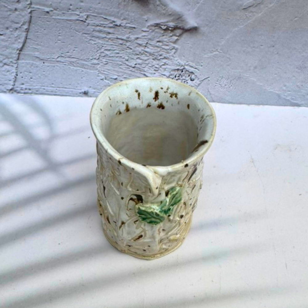 A handmade ceramic vase with a textured, organic shape, featuring a cream-colored glaze with brown speckles, a small green leaf detail on the front, and a small pouring spout, set against a white surface and textured gray wall.