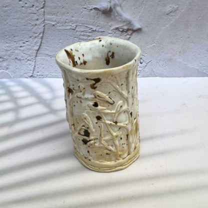 A handmade ceramic vase with a textured, organic shape and a cream-colored glaze speckled with dark brown, set against a white surface with soft window shadow patterns.