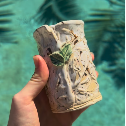 A hand holding a small handmade ceramic bud vase with a textured tree-like relief and a green leaf detail, set against a blurred turquoise water background.