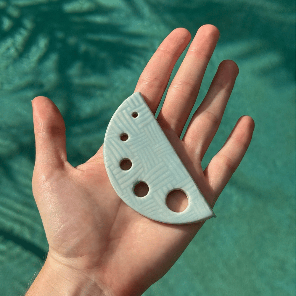Hand holding a ceramic half-circle herb shredder with three holes against a teal background