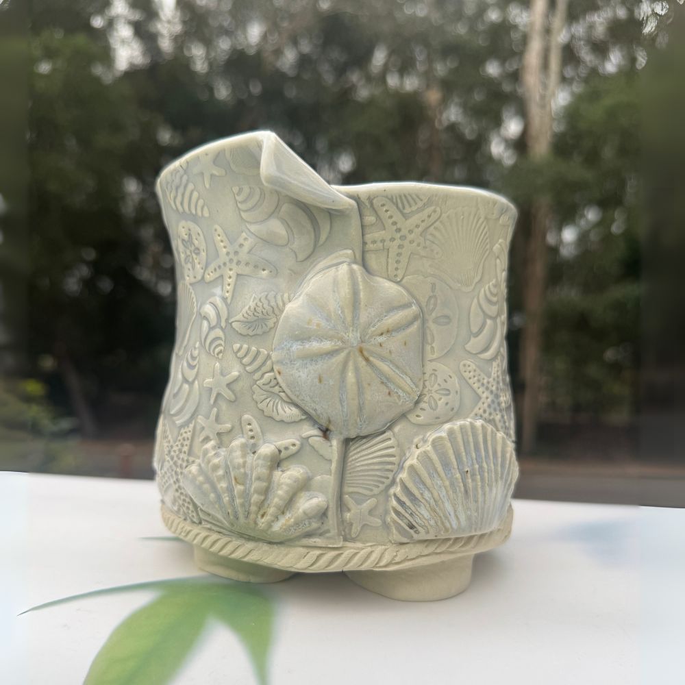 Ceramic planter with sand dollar and shell design on a white surface with blurred greenery in the background