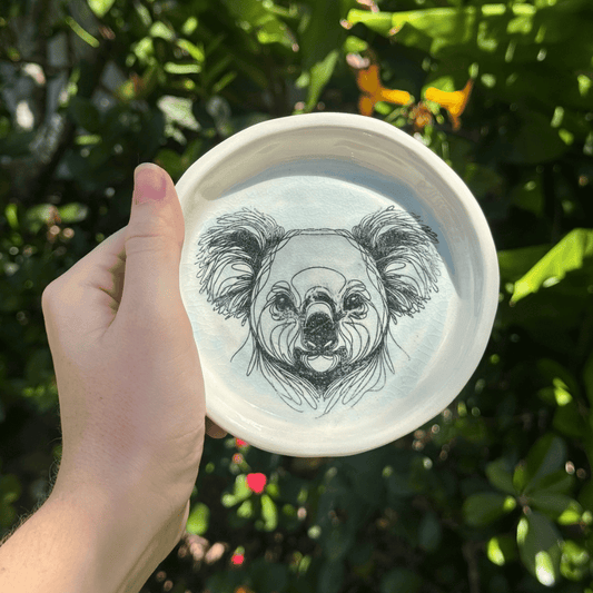 Hand holding a white dish with a black and white illustration of a koala on a green leafy background
