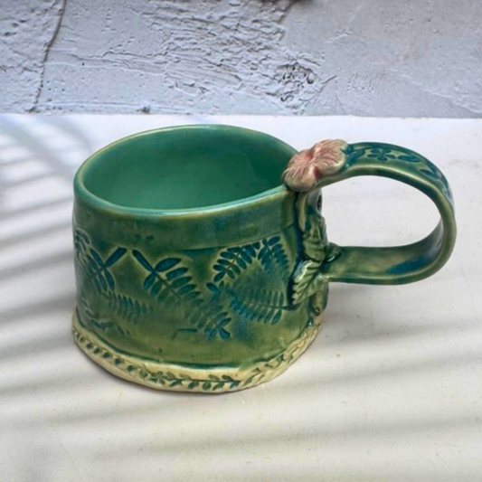 A green glazed ceramic mug with embossed leaf patterns and a small pink flower accent on the handle, set against a textured white wall with soft shadow overlays.