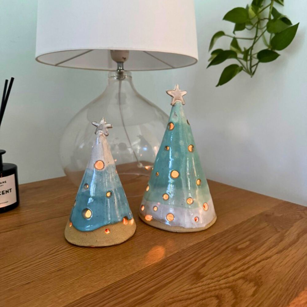 Two ceramic Christmas tree luminary cones on a wooden surface with a lamp in the background.