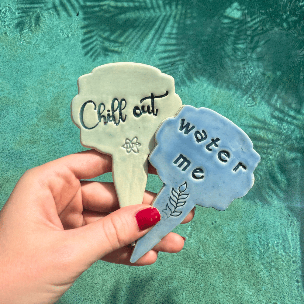 Hand holding two plant markers with 'Chill out' and 'Water me' text against a teal background. Australian Pottery.