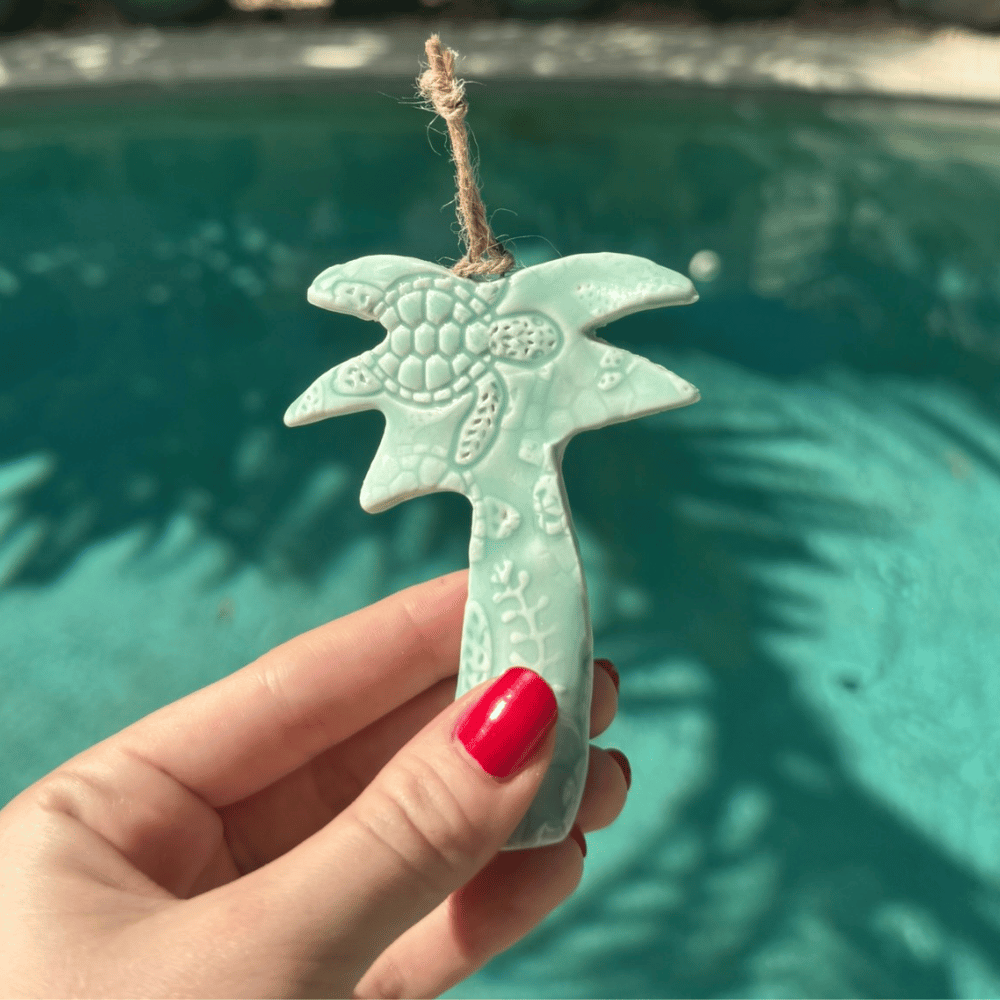 Ceramic palm tree ornament held by a hand with red nail polish against a pool background. Sunshine Pot-Tree, Sunshine Coast, Qld