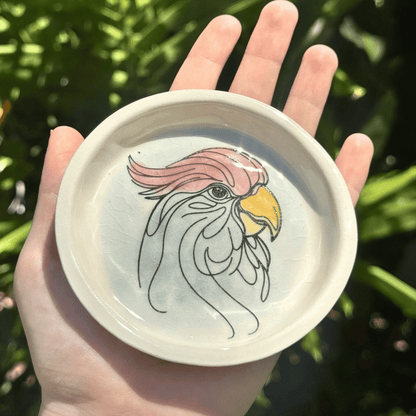 Hand holding a small round ring dish with a cockatoo illustration against a green leafy background